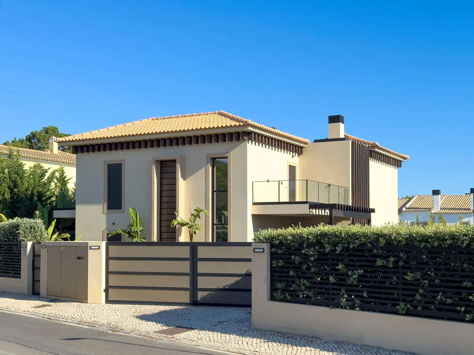 Two-storey Algarve villa with a terracotta roof, slatted gates and a clipped hedge under a clear sky